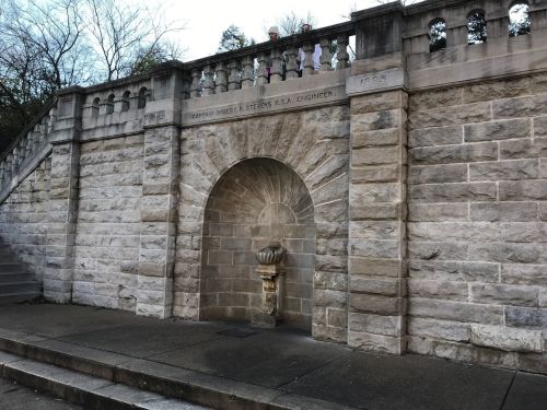 Stone wall with an arched entrance and decorative urn, featuring a staircase and railing above.