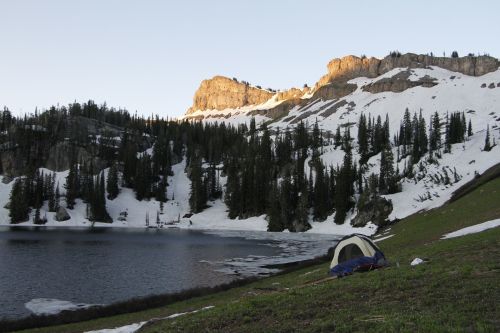 A tent set up by a snowy lake, surrounded by mountains and pine trees under a clear sky.