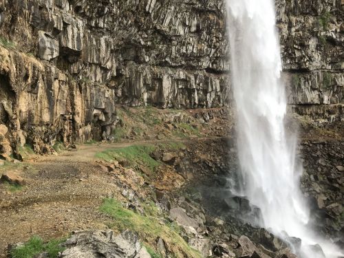 A waterfall cascades down rocky cliffs, surrounded by lush greenery and a gravel path.