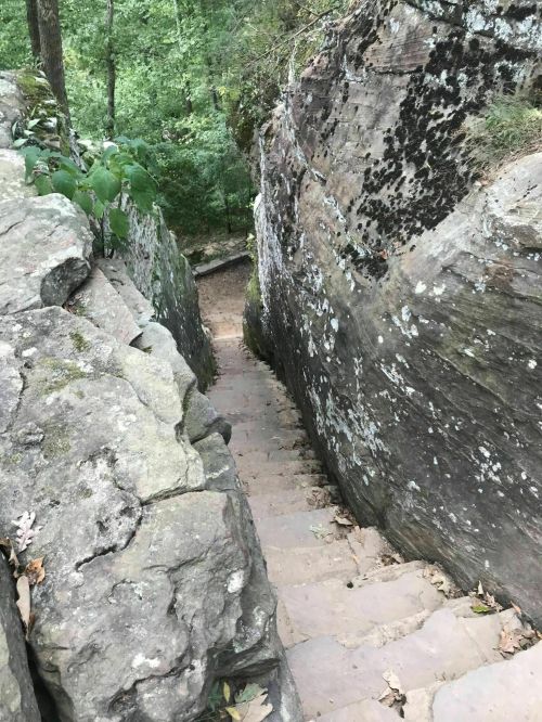 A narrow stone staircase winds between large moss-covered rocks in a wooded area.