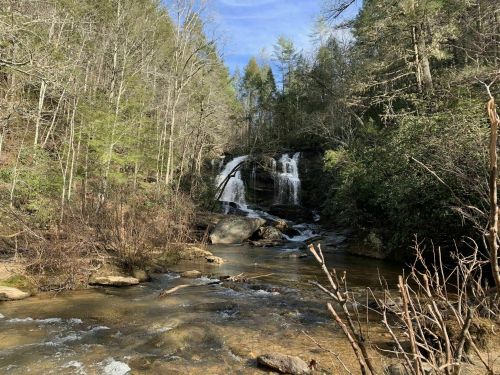 A serene waterfall cascades over rocks, surrounded by lush greenery and trees under a clear blue sky.