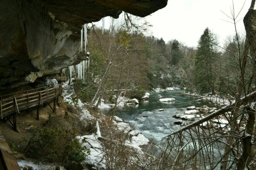 A winter landscape featuring a river, snow-covered trees, and icicles hanging from a rocky overhang.