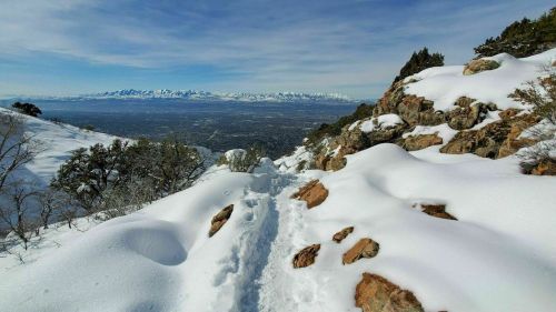 A snowy mountain trail winds through rocky terrain, with distant snow-capped peaks under a clear blue sky.