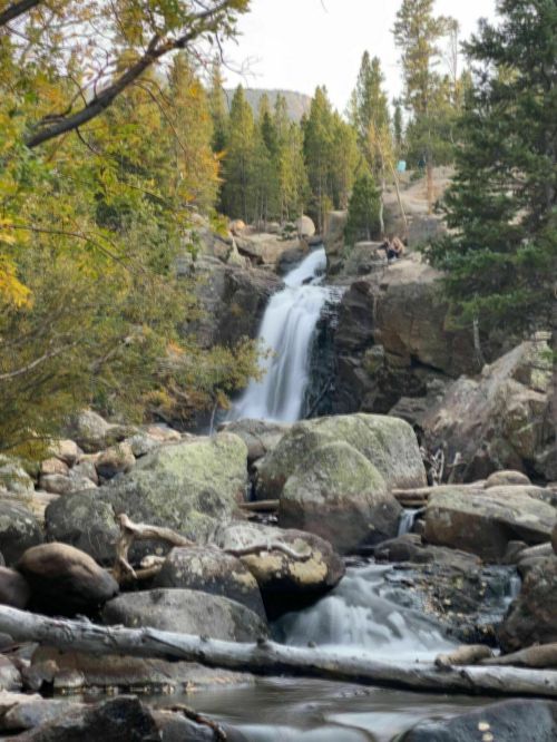 A serene waterfall cascades over rocks, surrounded by trees in autumn colors, with two people sitting nearby.