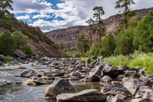 A serene river flows through a rocky landscape, surrounded by lush greenery and towering cliffs under a partly cloudy sky.