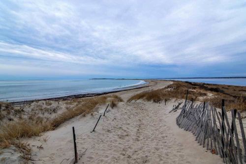 A sandy path leads to a tranquil beach, bordered by grass and wooden fencing, under a cloudy sky.