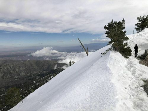 A hiker walks along a snowy ridge with mountains and clouds in the background under a cloudy sky.
