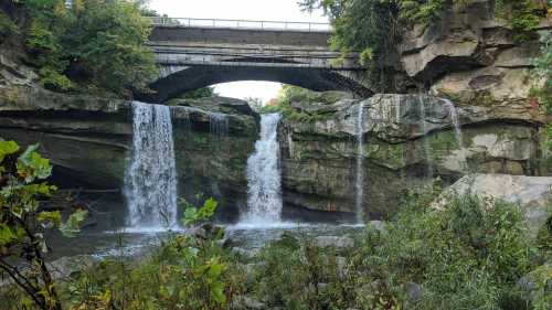 A scenic view of two waterfalls cascading over rocky cliffs, with a bridge overhead and lush greenery surrounding the area.