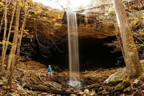A person stands near a waterfall cascading from a rocky overhang, surrounded by autumn foliage and trees.
