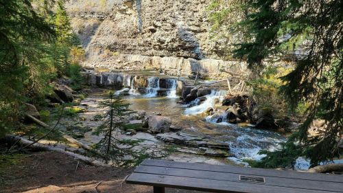 A serene river with small waterfalls, surrounded by rocky cliffs and lush greenery, with a wooden bench in the foreground.
