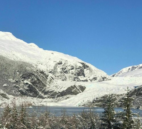 Snow-covered mountains rise above a serene lake, framed by evergreen trees under a clear blue sky.