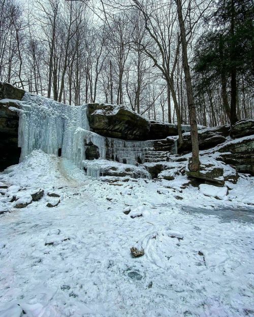 A frozen waterfall surrounded by snow-covered rocks and bare trees in a winter landscape.