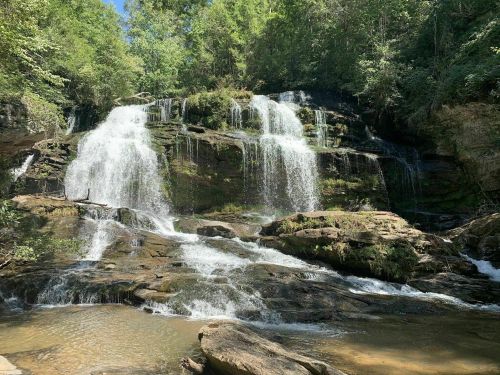 A serene waterfall cascading over rocky terrain, surrounded by lush green trees and a clear blue sky.
