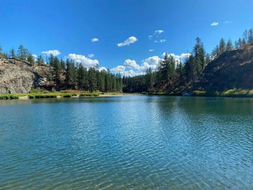 A serene lake surrounded by trees and rocky hills under a clear blue sky with fluffy clouds.