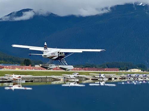 A seaplane takes off from a calm lake, reflecting mountains and a small airport in the background.