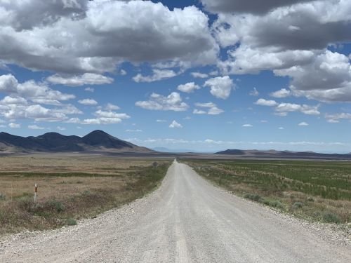 A gravel road stretches into the distance, flanked by fields and mountains under a blue sky with fluffy clouds.