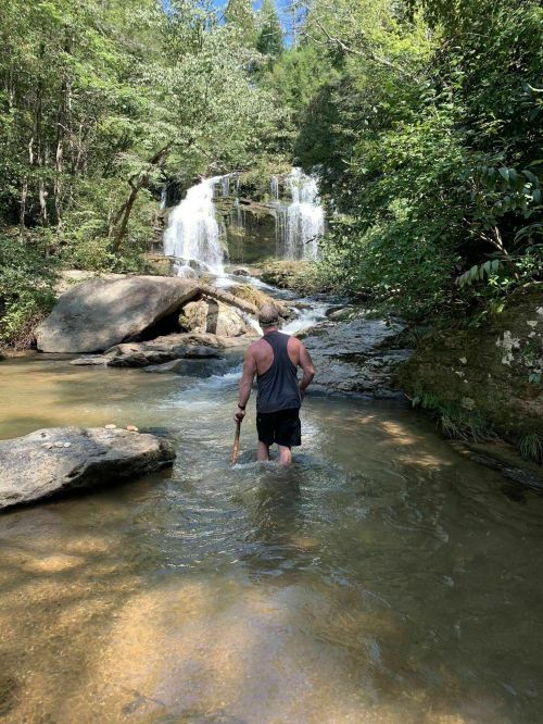 A person wades through a stream near a waterfall, surrounded by lush greenery and rocky terrain.
