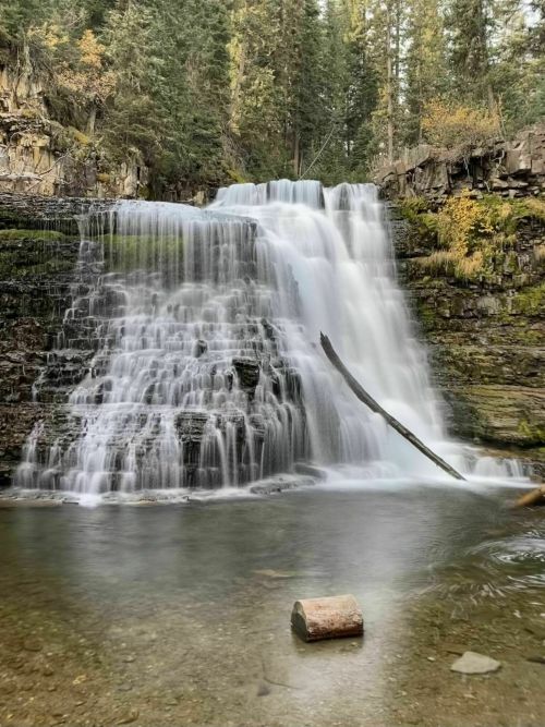 A serene waterfall cascades over rocky cliffs into a clear pool, surrounded by lush green trees and autumn foliage.