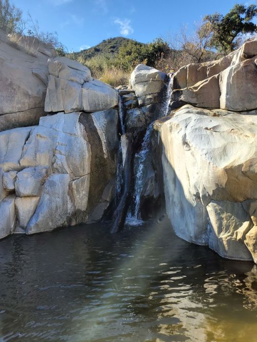 A small waterfall cascades over rocky cliffs into a serene pool, surrounded by greenery and a clear blue sky.