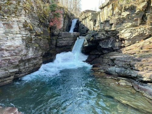 A serene waterfall cascades between rocky cliffs into a clear blue pool, surrounded by trees and natural beauty.