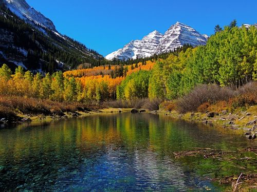 A serene mountain landscape with a clear lake reflecting vibrant autumn foliage and snow-capped peaks under a blue sky.