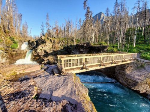 A wooden bridge spans a river near a waterfall, surrounded by rocky terrain and trees in a mountainous landscape.