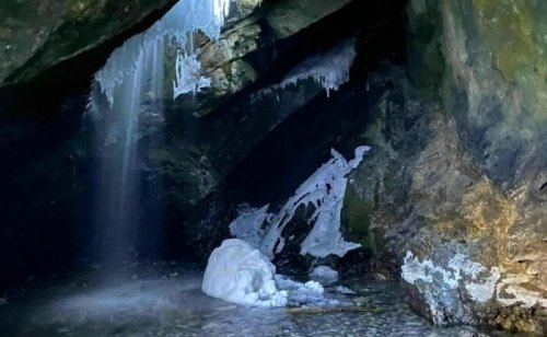 A dark cave with icicles hanging from the ceiling and a small waterfall flowing into a rocky pool.