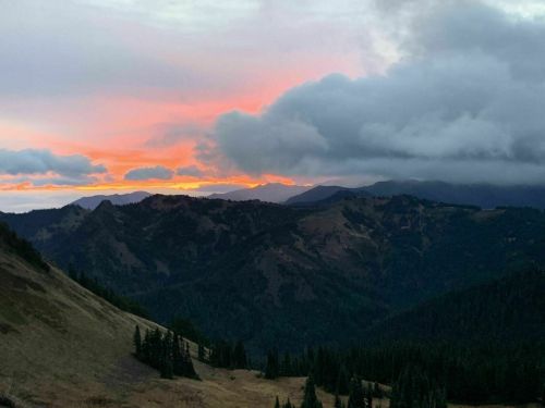 A scenic view of mountains at sunset, with vibrant orange and pink hues in the sky and dark clouds above.
