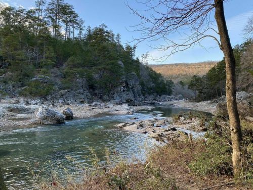 A serene river flows through a rocky landscape, surrounded by trees and hills under a clear blue sky.