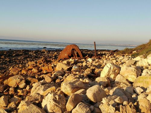 Rusty shipwreck on a rocky beach with calm water and a clear sky in the background.