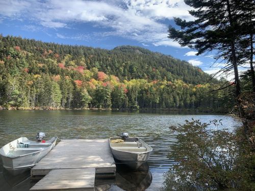 A serene lake surrounded by colorful autumn trees, with two boats docked at a wooden pier under a blue sky.