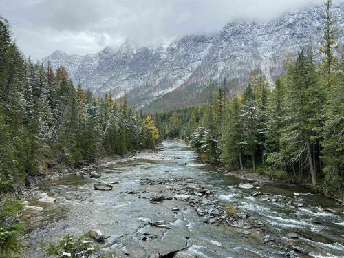A serene river flows through a forested valley, surrounded by snow-capped mountains under a cloudy sky.