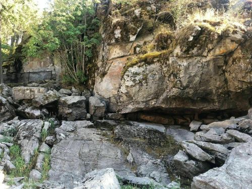 Rocky cliffside with moss and greenery, featuring large boulders and a small water source at the base.