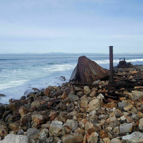 A weathered shipwreck rests on rocky shorelines, with waves crashing against the stones under a cloudy sky.