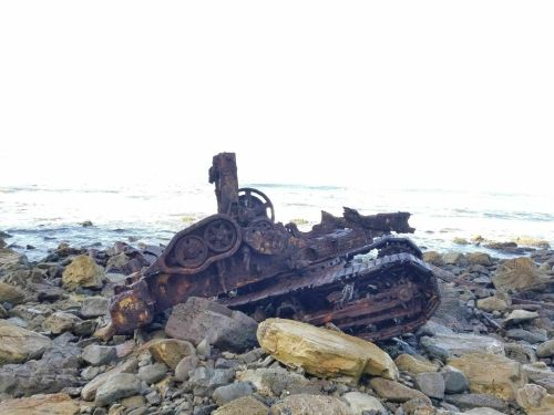 Rusty tank wreckage on a rocky beach, partially submerged near the water's edge.