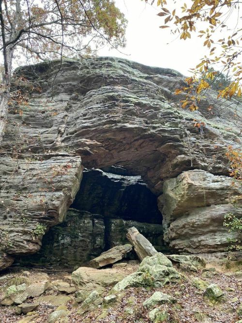 A rocky cave entrance surrounded by trees and autumn foliage, with large boulders in the foreground.