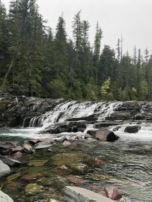 A serene waterfall cascades over rocky ledges, surrounded by lush green trees and a clear, flowing river.