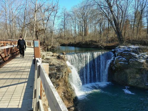 A person walks along a wooden bridge beside a waterfall surrounded by trees in a serene natural setting.