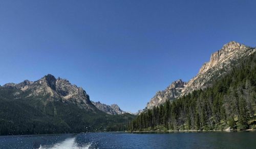 A serene lake surrounded by mountains and dense forests under a clear blue sky. Water splashes in the foreground.