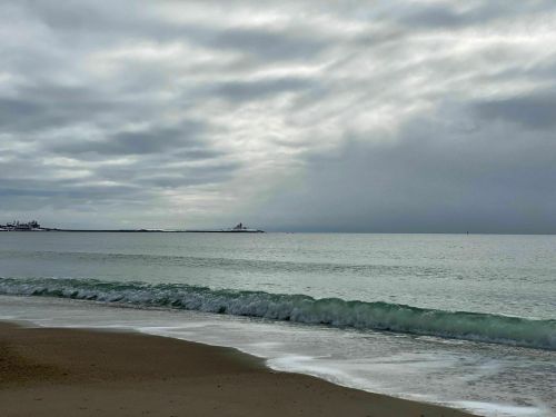 A calm beach scene with gentle waves, overcast skies, and a distant lighthouse on the horizon.