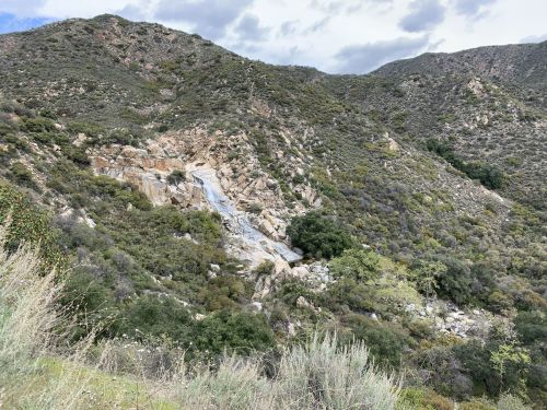 A rocky hillside with a small waterfall cascading down, surrounded by green shrubs and trees under a cloudy sky.
