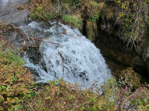 A small waterfall cascades over rocks, surrounded by greenery and autumn foliage.