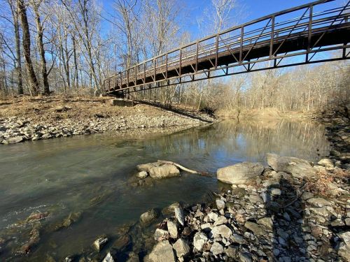 A metal bridge spans a calm river, surrounded by bare trees and rocky banks under a clear blue sky.