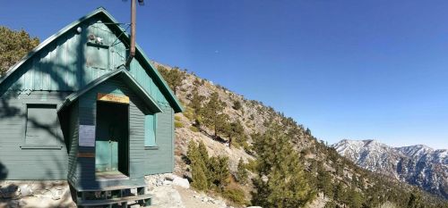 A green cabin sits on a mountainside, surrounded by trees and snow-capped peaks under a clear blue sky.