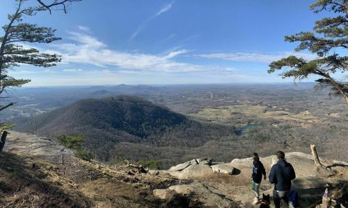 A panoramic view from a mountain peak, showing rolling hills and valleys under a clear blue sky. Two hikers are in the foreground.