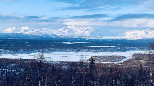 Snow-capped mountains rise above a frozen river, surrounded by a forest under a blue sky with scattered clouds.