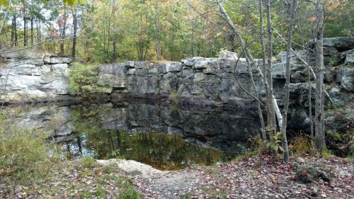 A serene pond surrounded by rocky cliffs and autumn foliage, reflecting the trees and sky above.