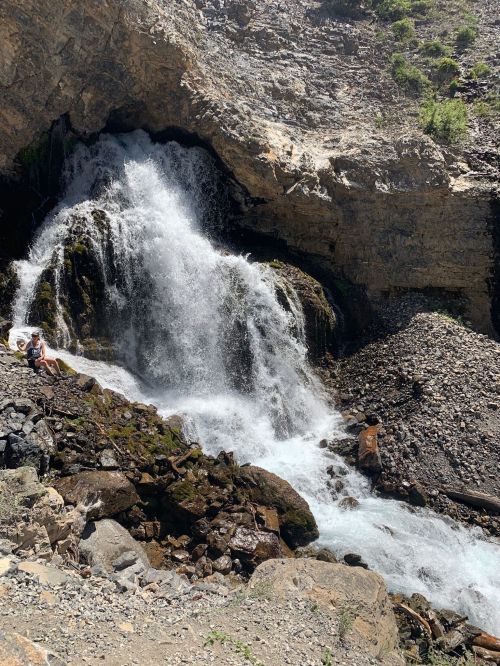 A waterfall cascades down rocky terrain, surrounded by boulders and greenery, with a person sitting nearby.