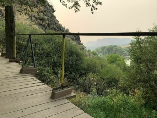 A wooden walkway with a railing overlooks a river surrounded by lush greenery and distant hills in a hazy atmosphere.