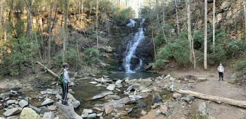 Two people stand near a waterfall surrounded by rocks and trees in a serene natural setting.